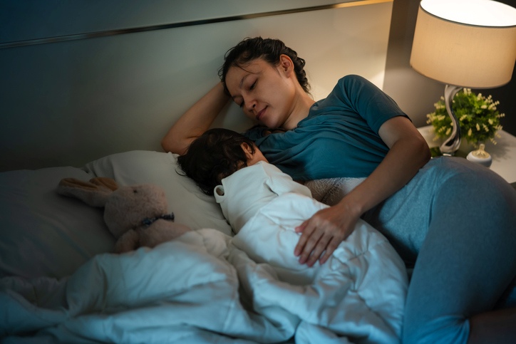 A parent resting beside a sleeping young child in bed