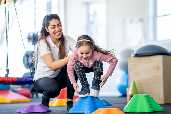 A therapist smiling at a young girl as she steps onto colorful balance cones in a therapy gym